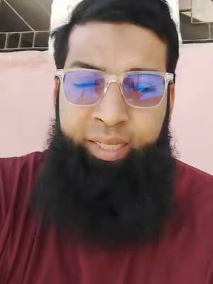 A selfie-style shot featuring a man with a dark beard and glasses in a red shirt against a light pink background. The man is looking directly at the camera.
