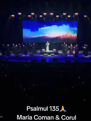 A live concert scene is captured in a vertical shot. The focus is on a stage with a large screen displaying a scenic background of a sky and fields. A woman in a light-colored gown is centered on stage, singing with a choir behind her, dressed in dark attire. The stage is illuminated with blue and white lights. Text appears at the bottom: 'Psalmul 135' and 'Maria Coman & Corul'.