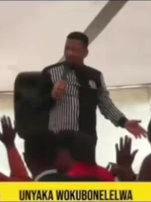 A man with short dark hair is speaking into a microphone. He's wearing a black and white striped jacket. In the foreground, there are raised hands of the audience. A yellow banner at the bottom with text in a language I don't recognize.