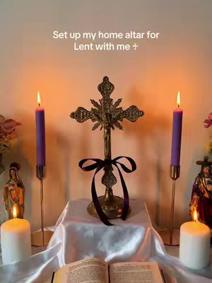 A photograph shows a home altar set up for the Lenten season. A golden cross with a black ribbon is centered on a white cloth. Two purple candles in gold holders flank the cross, and in front of the cross is an open book. Two white candles and two small statues are visible on either side