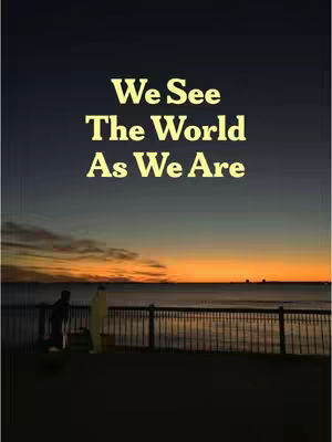The image portrays a sunset scene over a body of water, with a silhouetted cityscape visible on the horizon. In the foreground, two figures stand on a structure, appearing as dark silhouettes. The sky transitions from deep blue to orange and yellow, creating a warm, serene atmosphere, and the text overlay reads 'We See The World As We Are'.