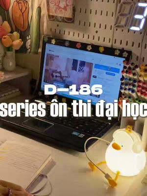 The cover image shows a study setup with a laptop as the focal point, displaying a video interface. The laptop sits on a desk with a notebook, a duck-shaped lamp, a digital clock displaying the number 3, and colorful pens nearby. The background includes some decorative elements such as a wall decorated with small items, and a flower vase.