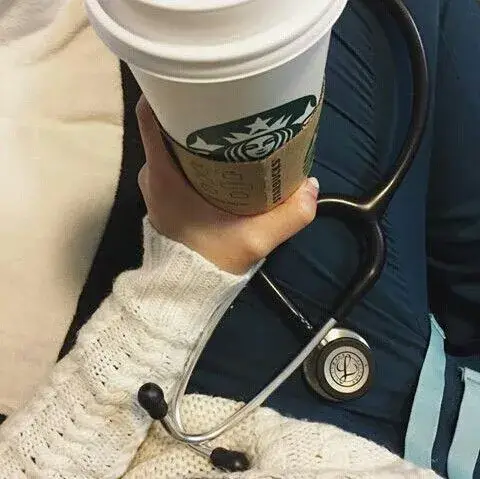 A hand wearing a white sweater, holding up a Starbucks coffee cup with a stethoscope draped around it. The cup is positioned at an angle, partially obscuring the person's upper body. The stethoscope's earpieces are black, contrasted against the cup's white and the person's fair skin. The photo is well-lit, showcasing a close-up view of the scene.