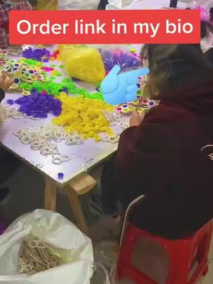 The cover image shows a behind-the-scenes view of a workspace filled with colorful pop-it toys. A person is partially visible, seated at a table and seemingly assembling or organizing the toys. Overhead, a red banner displays the text 'Order link in my bio.'