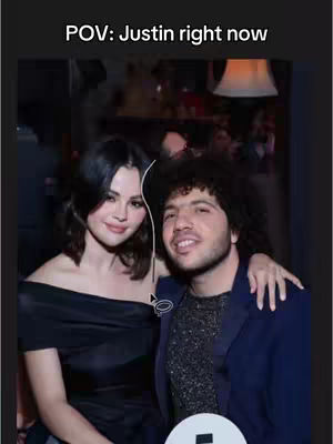 A candid, somewhat blurry image of a social setting. It features two people close together, a woman on the left, and a man to the right. The woman is wearing an off-the-shoulder black dress, looking at the camera. The man has an arm around her shoulders. The overall scene suggests a social gathering at night, possibly at a club.