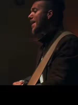 The image features a man from the side with his mouth open as if singing or speaking. He wears a black jacket over a white shirt, and the strap of a guitar or instrument is visible. The background is dark, with warmer tones towards the top.