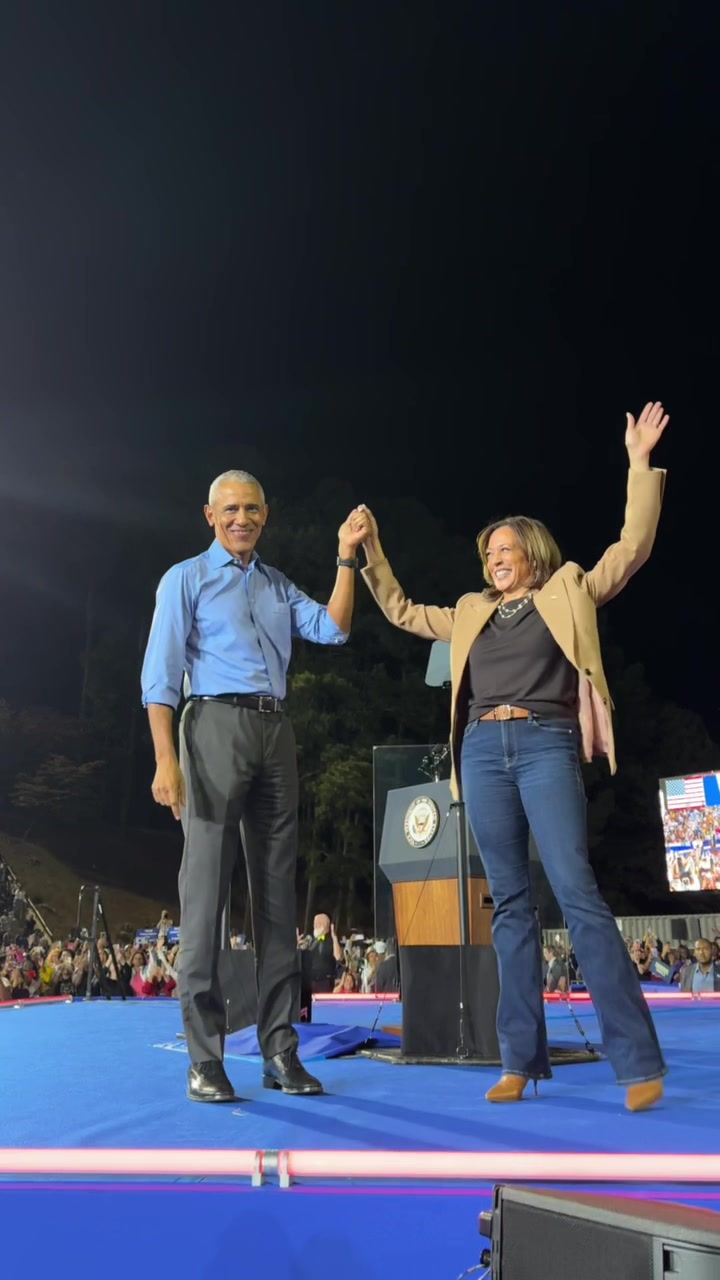 The image shows Barack Obama and Kamala Harris on a stage at night, holding hands and celebrating. Obama, on the left, wears a blue shirt and dark pants. Harris, on the right, is in a tan jacket, black top, and jeans, with a podium behind her. The background includes a crowd and some greenery, with a dark night sky overhead.