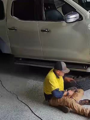 A man in a yellow shirt and baseball cap is kneeling in front of a beige pickup truck. He appears to be working on or inspecting something near the tire, with tools and equipment visible. The setting seems to be a garage or outdoor work area.