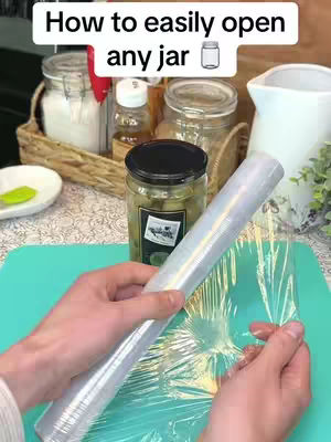 The image shows a close-up of a person's hands holding plastic wrap, alongside a teal cutting board and various kitchen items. In the background, jars and other kitchen supplies are visible. The setup suggests a how-to or tips-and-tricks video.