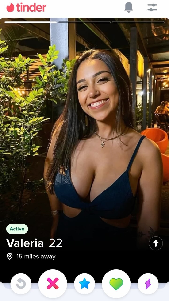A selfie from the Tinder app showing a young woman with long dark hair smiling at the camera. She is wearing a low-cut blue top. The background is blurred, possibly an outdoor restaurant setting. The Tinder interface displays her name, age, and distance from the viewer. The overall composition is bright and casual.