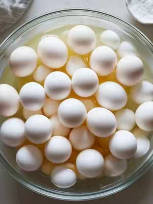 A close-up, overhead shot of a glass bowl filled with eggs, almost completely submerged in a yellowish liquid. The bowl is surrounded by a white surface, possibly a countertop, and there's a glimpse of a white cloth and another small white bowl in the upper right corner. The eggs occupy most of the frame, rounded and white, creating a dense pattern. The lighting is soft and highlights the textures of the eggs, the liquid, and the glass.