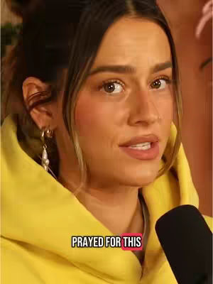 A woman with light brown hair is centered in the frame, looking slightly to the side as if speaking. She wears a yellow hooded sweatshirt and hoop earrings with dangling pearls. Behind her, the background is blurred, suggesting an indoor setting. A microphone is visible to her right, and a text overlay reading "PRAYED FOR THIS" is at the bottom, enclosed in a pink box.