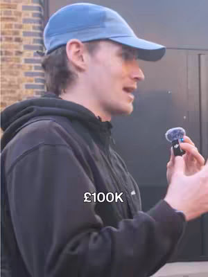 A man wearing a blue baseball cap and a black hoodie is talking into a microphone with a furry windscreen. The text overlay says '£100K'. The background includes a building with brick and dark gray or black siding.