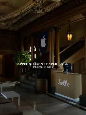 An interior shot of a grand building featuring a dark wooden staircase, ornate ceiling, and an Apple logo banner. In the foreground, there's a reception desk with the Apple logo and the word 'hello' on it.