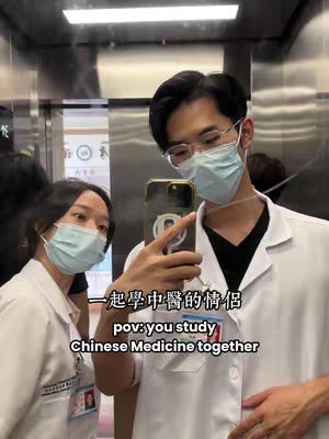 A selfie taken in an elevator shows a man and woman, both wearing medical masks and white coats. The man in the foreground holds the phone, capturing the image, with his face and attire clearly visible and partially obscuring the woman beside him. The elevator interior, with its metal doors, provides the background.