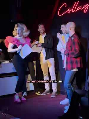 The image shows a group of people at a bar or club setting. A drag queen in a pink frilly top and black outfit stands holding a paper, with the mouth wide open in an expression of surprise. Other people (men) stand in the background, seemingly engaged in the event. The word "Collage" is in neon on the wall.