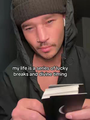A man with a slight smile looks down, holding a deck of cards. He wears a black beanie, a black hooded jacket, and an earring. Text overlayed on the image reads, 'my life is a series of lucky breaks and divine timing.'