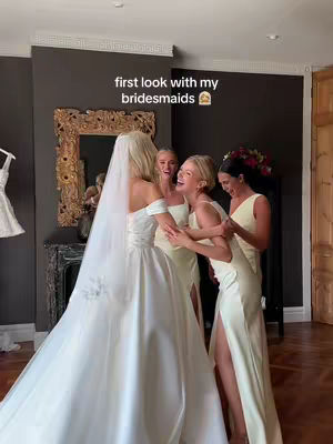 A bride in a wedding gown and veil is surrounded and embraced by four bridesmaids. The bride is in the center, facing the camera toward the left, with her bridesmaids on her right. The image is set in a well-decorated room with a large mirror.