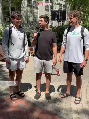 The image shows a street interview setup. Three young men, college students, are standing on a brick-paved area. A person in the middle is holding a microphone and speaking. The background shows a building with large windows. The scene is well-lit, suggesting an outdoor setting.