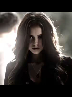 The cover image features a woman with long brown hair, looking directly at the viewer with a serious expression. The lighting is dramatic, partially obscuring the background, with emphasis on her face and upper body. She wears a dark top and a pendant necklace is visible. The overall composition is dark with a focus on her intense gaze.