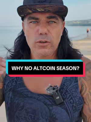 A man with long black hair and a black cap stands on a beach. He's wearing a dark blue tank top, and a small microphone can be seen attached to his shirt. The background shows a beach, the ocean, and some people in the distance.