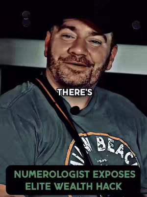 The cover image is a selfie-style shot. The visible subject is a man with fair skin, a beard, and short brown hair. He's wearing a dark baseball cap, a grey t-shirt with a graphic design and a black strap over his right shoulder. The image is well-lit suggesting a professional environment. The text overlay includes the words 'THERE'S' at the top and a lower rectangular block with 'NUMEROLOGIST EXPOSES ELITE WEALTH HACK'.