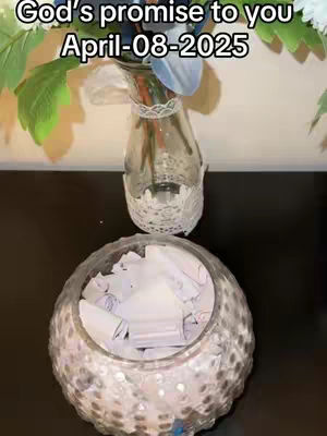 An overhead shot features a dark, glossy surface upon which two glass containers are placed. The top container is a vertically-oriented glass cylinder with a decorated rim, and it holds a few sprigs of green foliage. Below, sits a larger, round container filled with numerous rolled-up pieces of paper. Written text in the top left corner reads "God's promise to you April-08-2025".