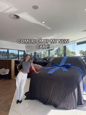 A woman stands in a car dealership, leaning against a car covered with a black sheet and a blue ribbon.