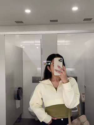 The image is a selfie taken inside a public restroom. A young woman is holding the camera in front of her face, obscuring most of her features. She is wearing a light beige zip-up sweater over a green top. The woman's dark hair, and part of her face, are visible. The background includes white walls and a closed stall door. Below the woman, a glimpse of black pants and a beige bag are visible. The top of the image captures the ceiling lights.