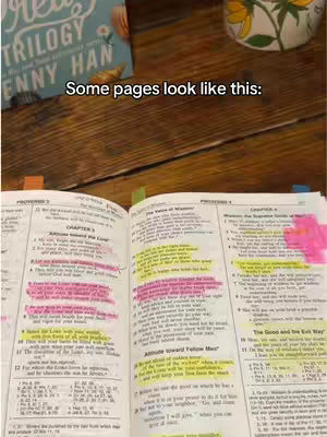 The image shows an open Bible on a wooden surface. The pages are marked with colorful highlighters and sticky notes. In the background, a book titled 'Rea Trilogy' is partly visible along with a decorative mug.