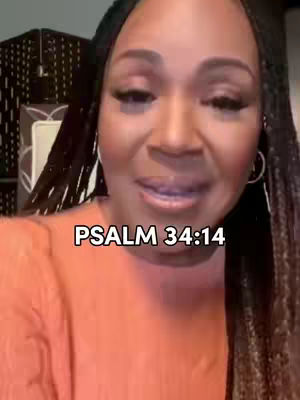 A portrait shot of a woman with dark curly braids, wearing a coral-colored top. The background is slightly blurred, suggesting an indoor setting. Text in a black box at the bottom reads, 'PSALM 34:14'.