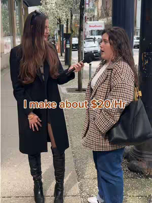 Two women stand on a sidewalk, one interviewing the other. The interviewer, wearing a black coat and boots, holds a microphone. The interviewee, wearing a plaid jacket and jeans, is the focus of the interview. The background shows a city street with buildings and parked cars.
