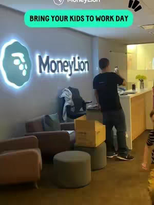 An office scene at MoneyLion featuring a man in a black shirt and jeans. The background features the MoneyLion logo, along with office furnishings like chairs, and a reception desk.
