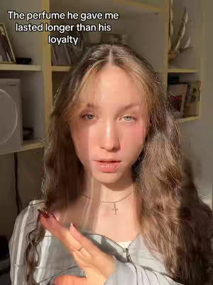 A young woman is centered in the frame, with her face taking up the majority of the space. She has long, wavy brown hair that frames her face, with slight highlighting. Her skin appears fair with some blush applied on her cheeks and lips. She is looking directly at the camera with a slightly serious expression. There is text overlaid on top. The lighting is soft and natural, suggesting a daytime setting. Behind her is a bookshelf.