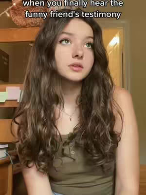 A young woman with long, wavy brown hair is centered in the frame, slightly angled towards the viewer with a contemplative expression. She wears a simple, sleeveless olive-green top, and a delicate necklace with a small pendant. The background is softly blurred, featuring glimpses of room elements. The image has a warm, natural lighting.