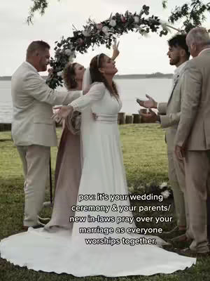 A wedding scene where a bride in a white dress is surrounded by wedding guests.  People are standing in the background on a green lawn.