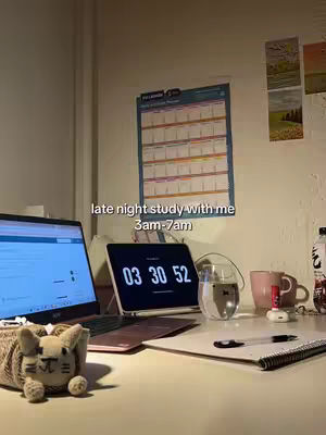 A well-lit desk setup with a laptop, tablet displaying a digital clock, a beverage, and study materials. There's a calendar on the wall, and a plush creature is near the laptop. The scene suggests studying late at night.