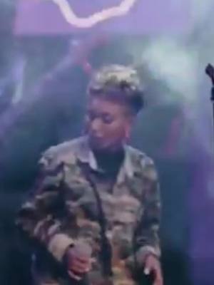 The cover image features a person on stage, captured in a mid-shot. The person, who appears to be a woman with short, styled hair. They are wearing a camouflage jacket. The background is blurred with colored stage lighting and fog effects.