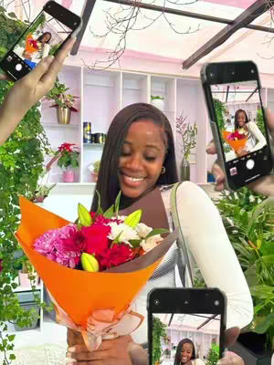 A smiling Black woman holding a bouquet of flowers in an orange wrapper. Around her are hands holding smartphones, each displaying a picture of her holding the flowers. The background shows a well-lit shop with plants and decorations.