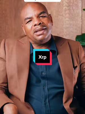 A close-up shot of a Black man in a brown suit jacket, blue shirt, and a black square with the word 'Xrp' in white text. The man is speaking, and behind him is a brown or tan wall and a bit of green foliage.