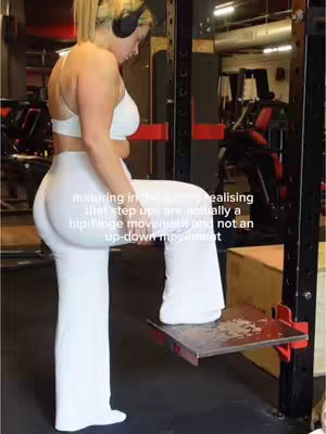 A woman in a gym is demonstrating a step-up exercise. She's wearing a white sports bra and matching white pants. She has blonde hair tied back and is wearing headphones. The background shows gym equipment.