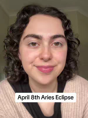 A close-up shot of a person with curly dark hair. The person is smiling slightly with pink lipstick. The text overlay at the bottom states “April 8th Aries Eclipse”. The background is a light beige color.