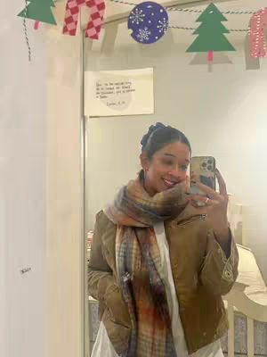 A woman is taking a selfie in front of a mirror. She's wearing a scarf and a jacket, and there are Christmas decorations hanging above.