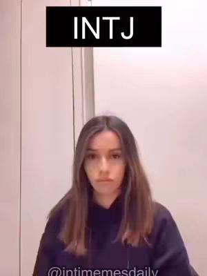 A woman faces the camera, with a neutral expression. She is wearing a dark hoodie. The background appears to be a plain wall. Above her head is a black box with white text that reads "INTJ".