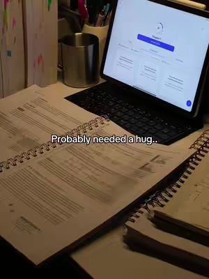 A desk is shown cluttered with study materials. On the desk, there's a partially open notebook with handwritten notes, some books, a mug, and a tablet displaying an online study platform. The lighting appears soft and warm, casting shadows on the items. Overlaid text reads 'Probably needed a hug..'.