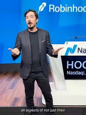 The cover image shows a man speaking at a podium, in front of the Robinhood logo. He has dark hair and is wearing a suit with his hands open gesturing outwards. The background is a bright blue screen that provides the Robinhood logo with white text. And behind him is a display screen with text. Text overlay is visible along the bottom edge of the image.