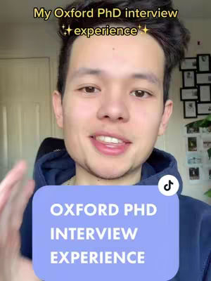 A young man with dark hair is centered in the frame of a selfie-style video. He's wearing a navy blue hoodie and smiling slightly, with a blurred background suggesting a room with a door and possibly some artwork. A large, light-blue rectangular graphic with white text stating "OXFORD PHD INTERVIEW EXPERIENCE" is overlaid on his chest, and a small TikTok logo is in the bottom right corner.