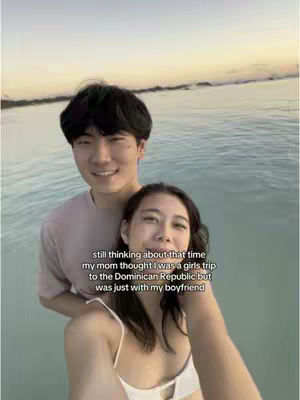 A selfie-style image of a couple waist-up in a body of water. The woman is in the foreground with her arms around the man, who is standing behind her. The background features water, a sunset sky, and a horizon line that indicates a beach or shore. Text is overlaid on the image.