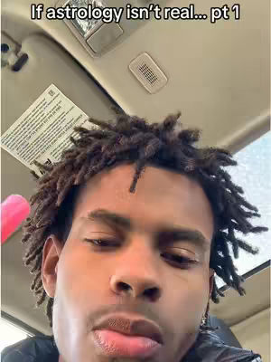 A selfie-style shot featuring a young Black man. His face is the primary focus, with his head angled slightly upwards and to the right. He has dark, closely-cropped hair. The lighting appears natural, possibly from overhead, casting soft shadows. The background shows the interior of a car setting, with a sun visor.