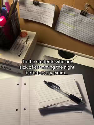 The image shows a close-up of a study setup. There is an open notebook with a pen on top, a book titled "James Clear", several pens and stationary items inside a pencil holder or cup, and notes and flashcards attached to the background with clips. The color palette is warm, dominated by the brown color of the surroundings.